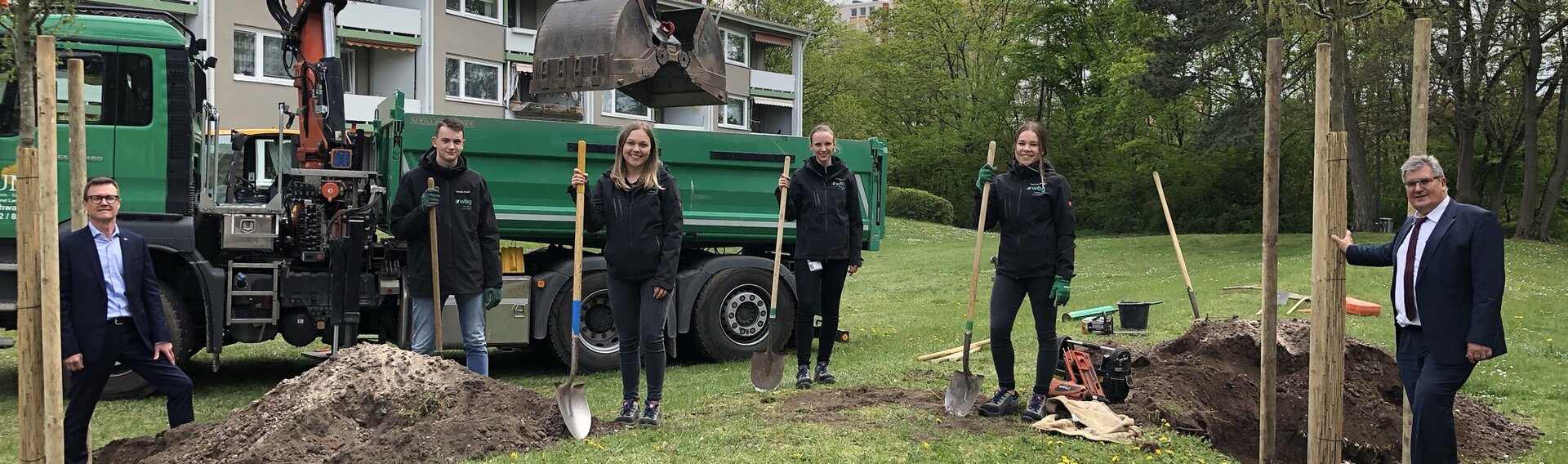 Five people are standing on a green area with freshly dug soil. On the left of the picture: wbg managing director Ralf Schekira. Next to him are four young people holding spades. A residential building and an excavator can be seen in the background.