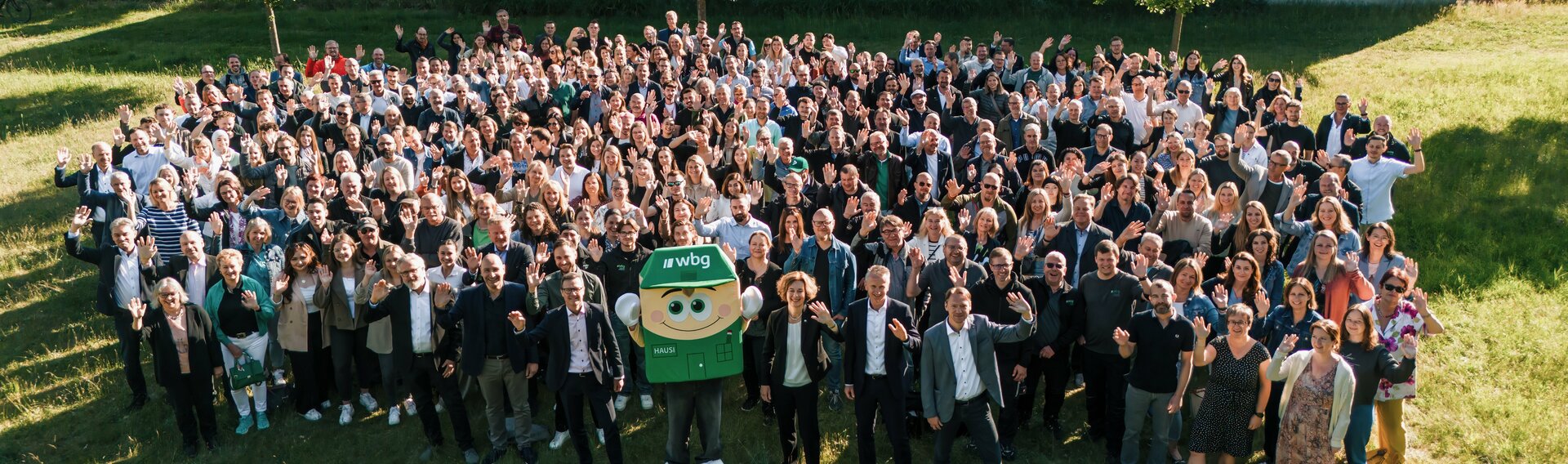 Eine große Gruppe von Menschen steht auf einer Wiese und winkt in die Kamera. Es sind Mitarbeiter der wbg Nürnberg. In der Mitte der ersten Reihe stehen der Geschäftsführer und die Bereichsleiter. Zwischen ihnen steht das Firmenmaskottchen „Hausi“. Es stellt ein grünes Haus mit einem Gesicht und Armen dar.
