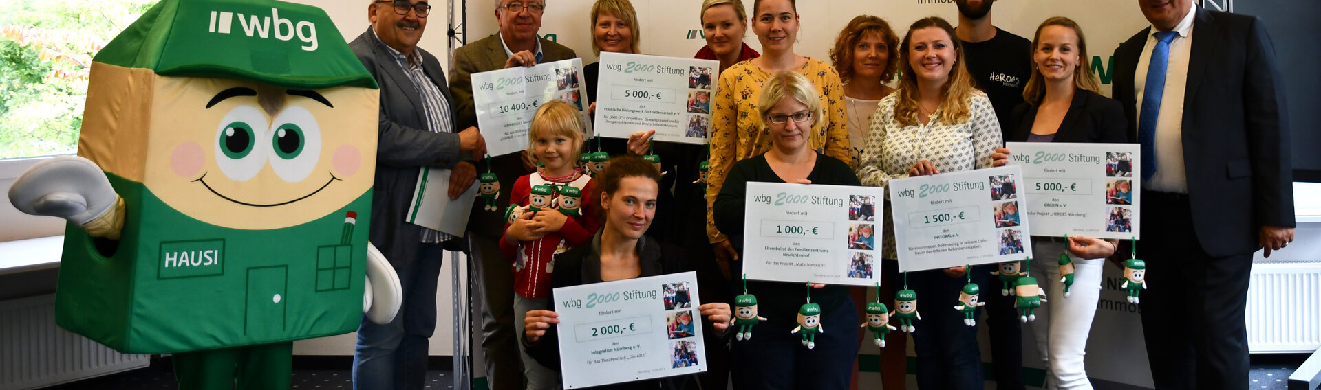 Mehrere Personen schauen in die Kamera und halten große, symbolische Spendenschecks der wbg 2000 Stiftung in den Händen. Links von Ihnen steht das wbg-Maskottchen „Hausi“. 