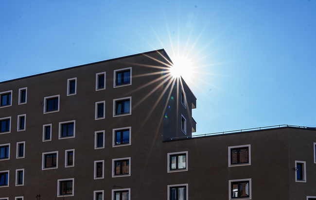 Beigefarbenes Gebäude mit vielen Fenstern. Die Sonne blitzt hinter der Fassade hervor.